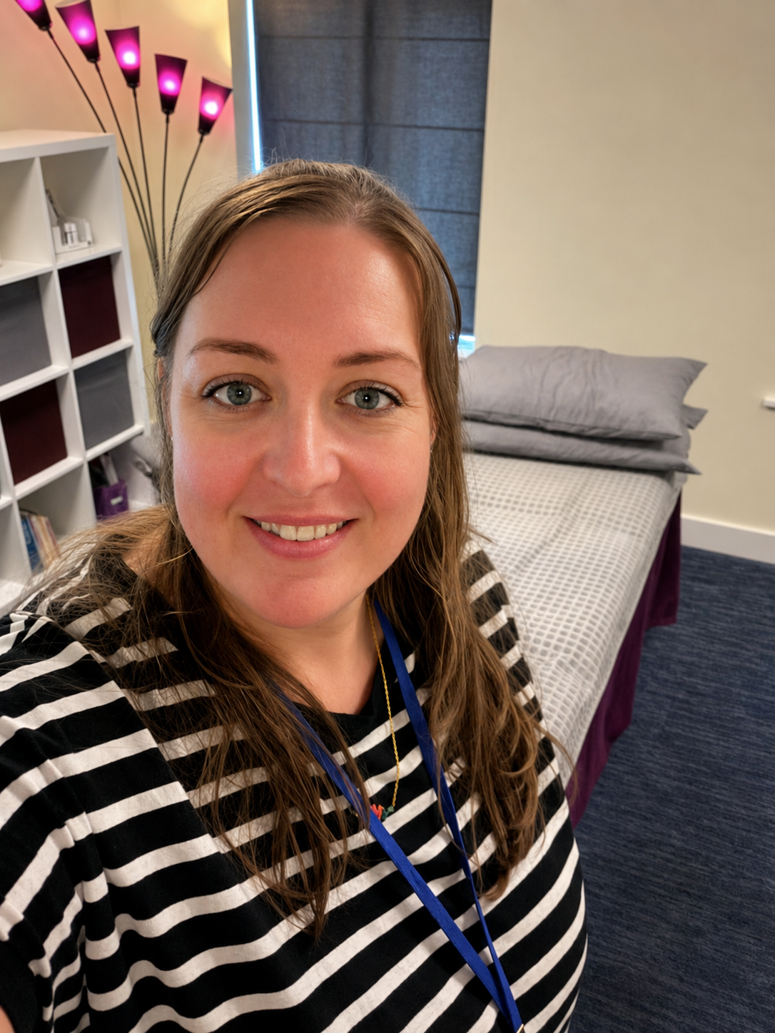 Smiling person in black-and-white striped shirt taking a selfie in a bedroom with a bed and floor lamp.