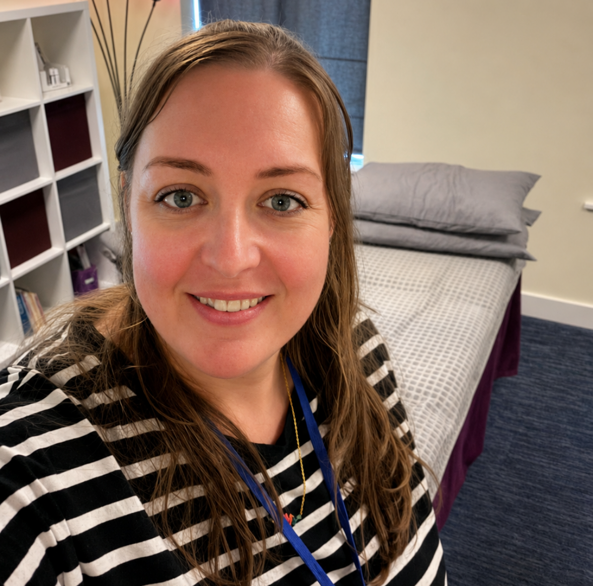 Smiling woman in striped shirt taking a selfie in a room with a bed and shelving unit.
