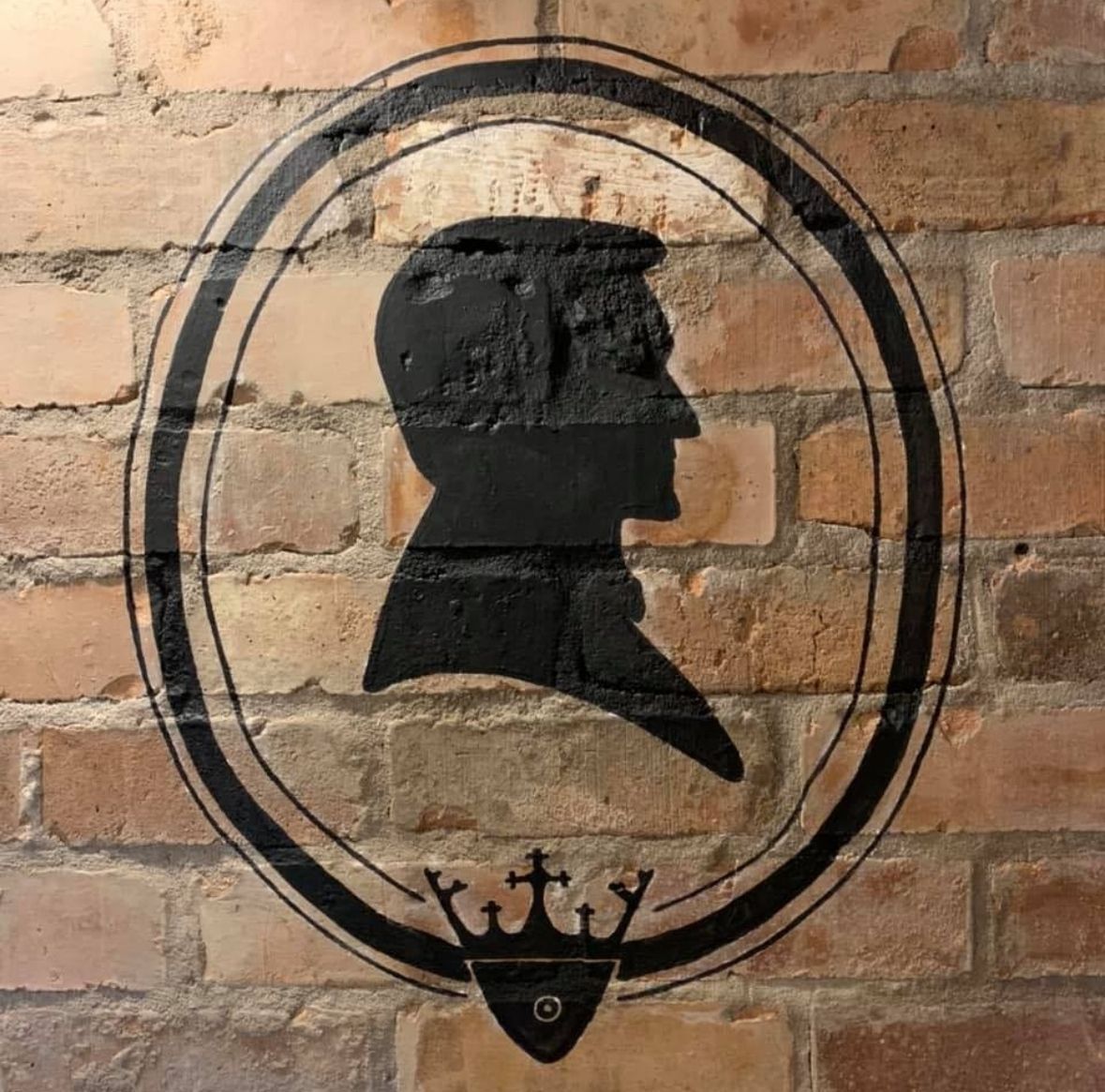 Black silhouette profile on an oval sign painted on a brick wall, with a small crown emblem below.