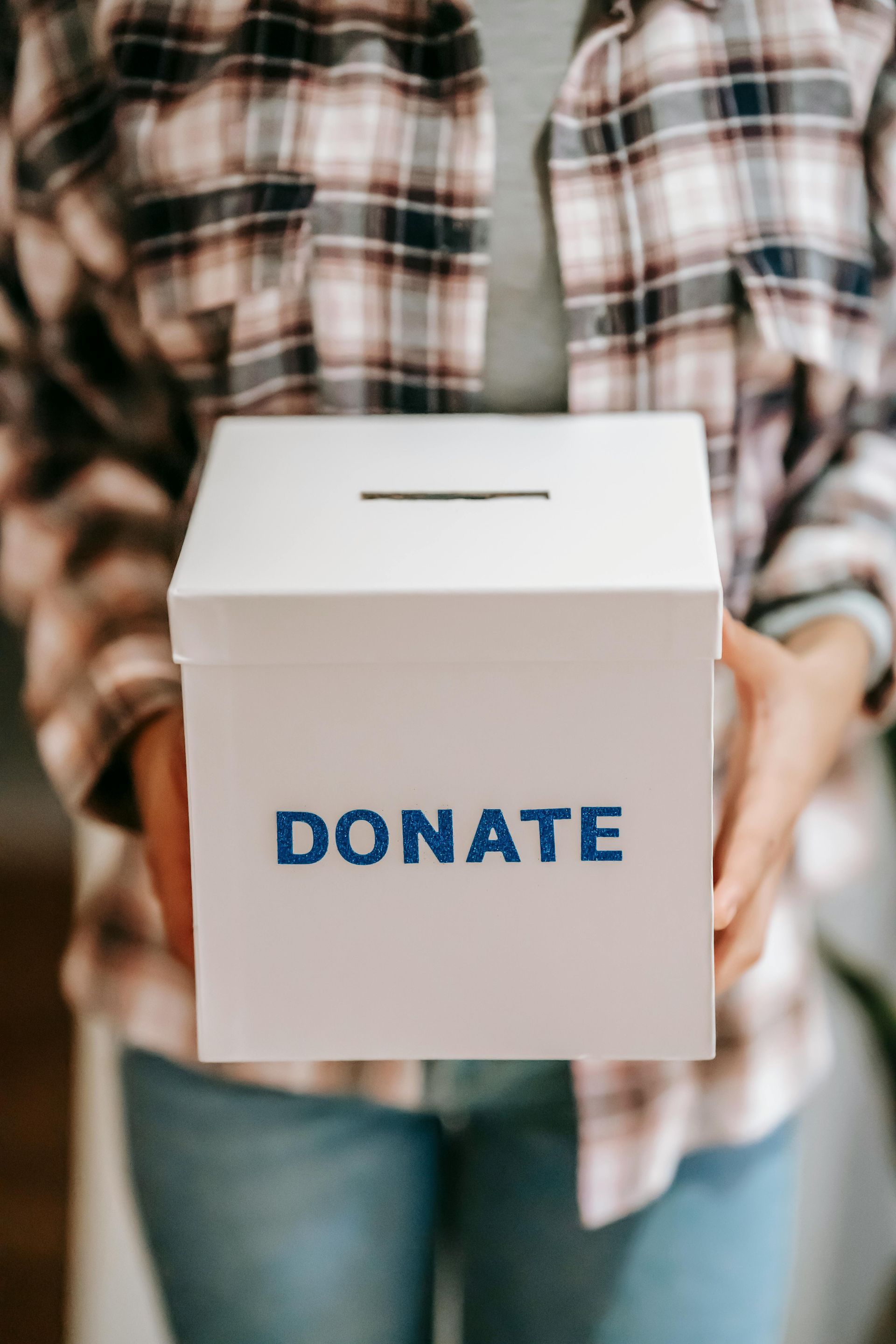 Person holding a white donation box labeled “DONATE” against a plaid shirt background