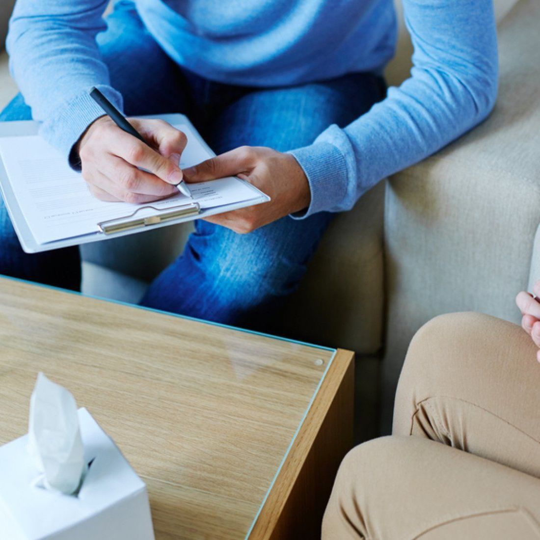 Person writing on a notepad at a table during a conversation; tissue box in foreground.