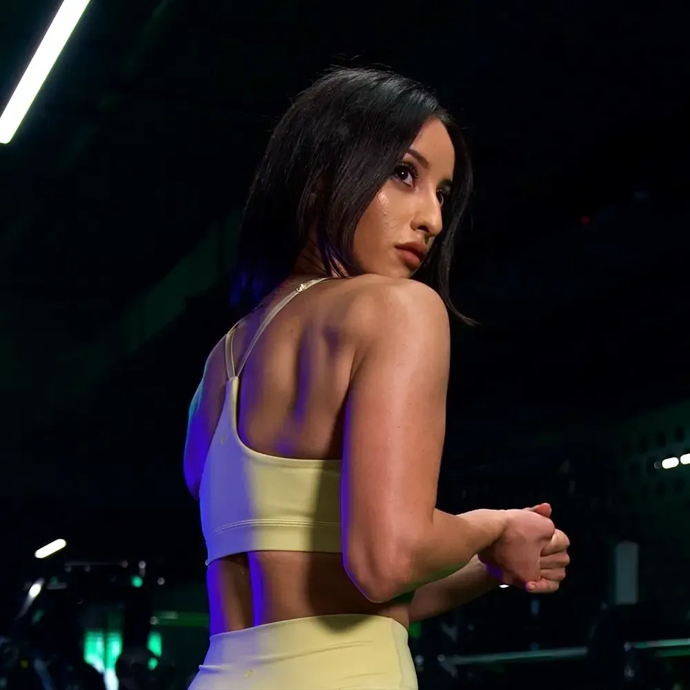 Leana in a yellow sports bra and leggings, looking back over her shoulder in a gym setting.