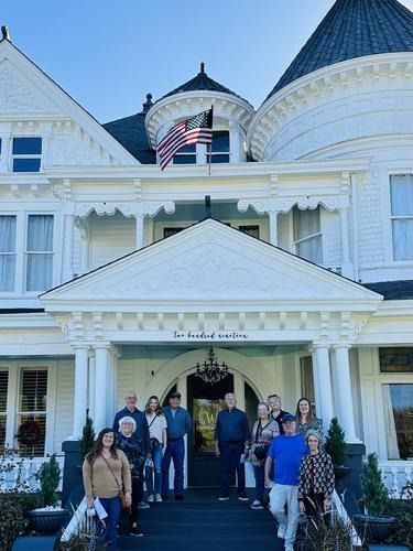 Group of people standing on the steps of a white Victorian house with an American flag.