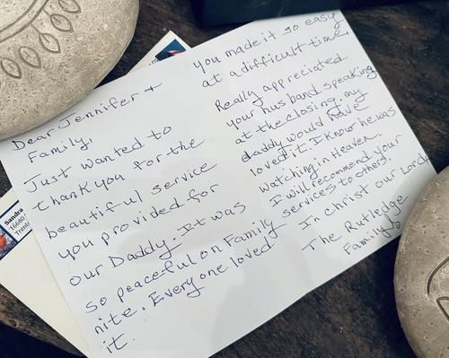 Handwritten thank-you note on a desk, with stones and an envelope. The note thanks the family for a service.