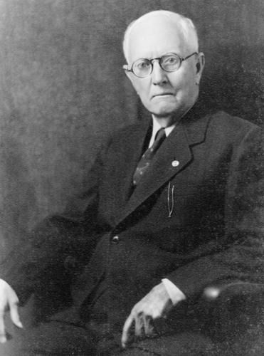 Elderly man in a suit, seated, wearing glasses, looking forward, serious expression.