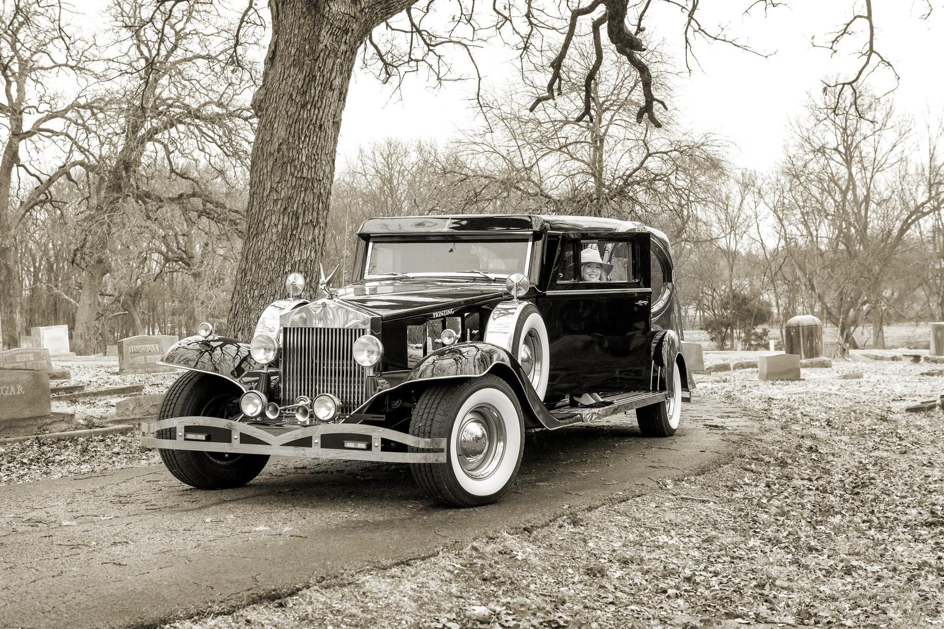 Classic black car with white tires parked in a cemetery on a brown path. Trees and headstones in the background.