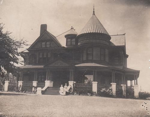 Large, dark Victorian house with a conical turret. People pose on the porch steps.