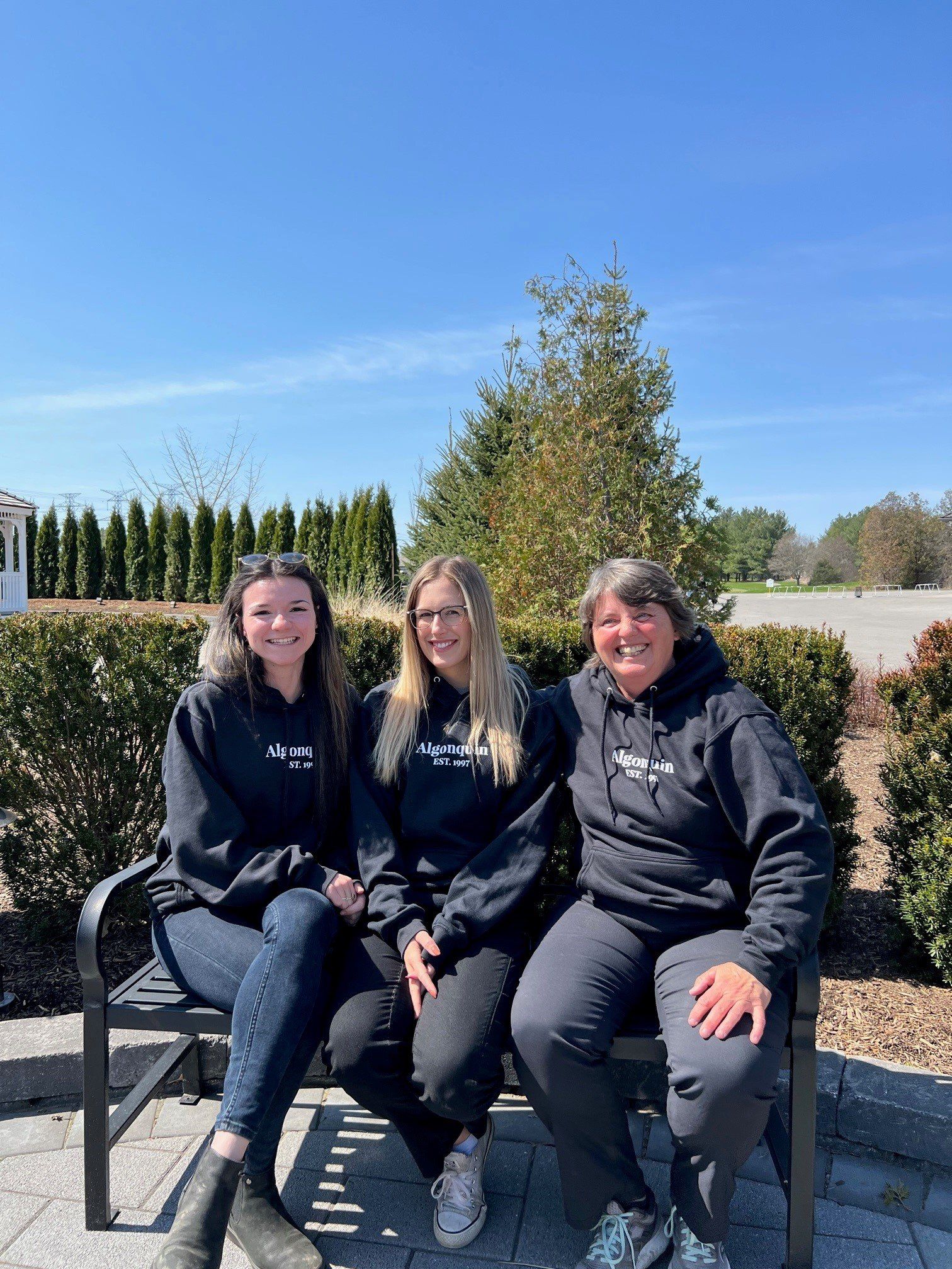 Three women are sitting on a bench wearing black hoodies.