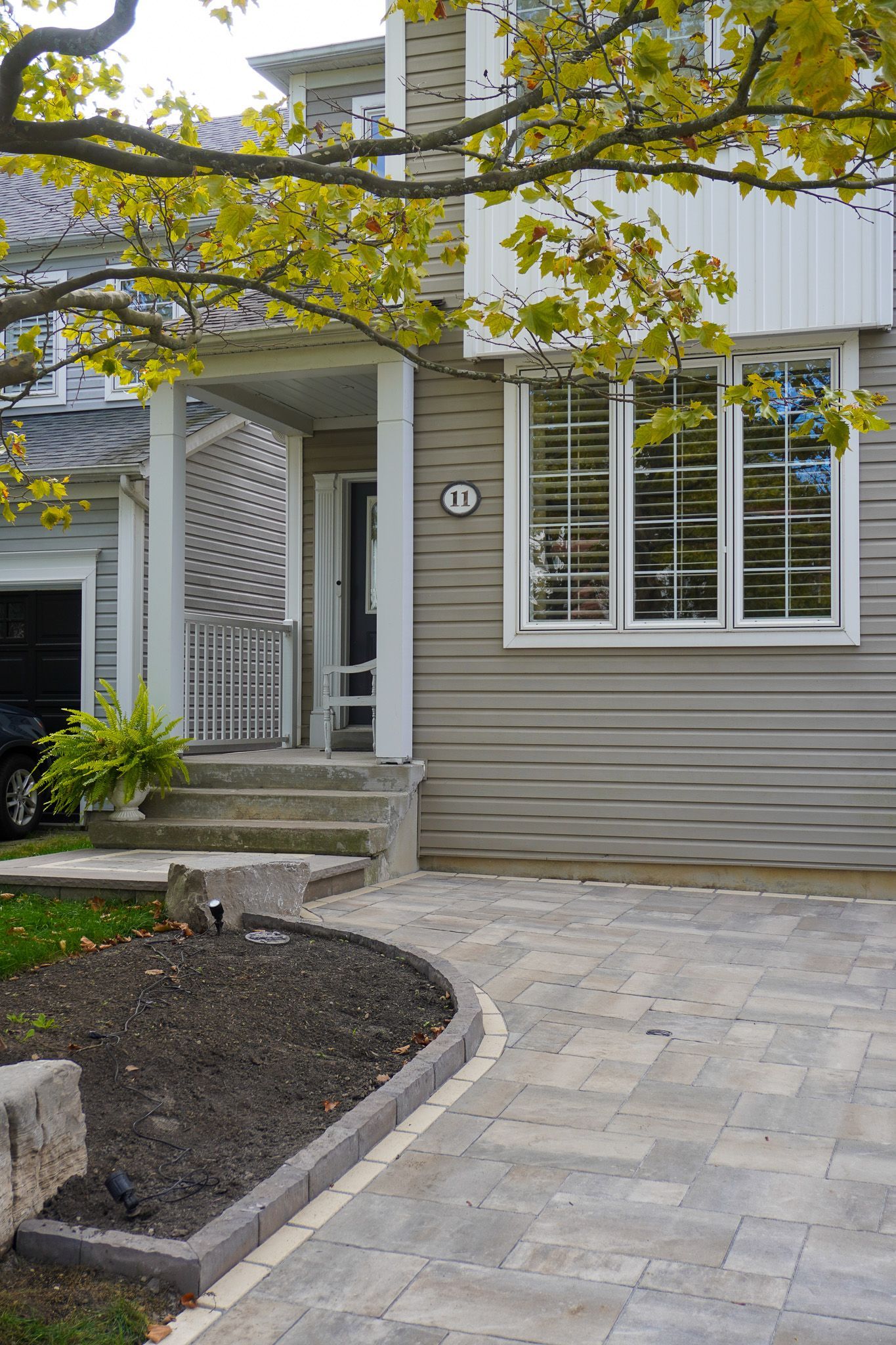 House exterior with paved walkway, porch, and window with shutters. Overhanging tree with yellow leaves.