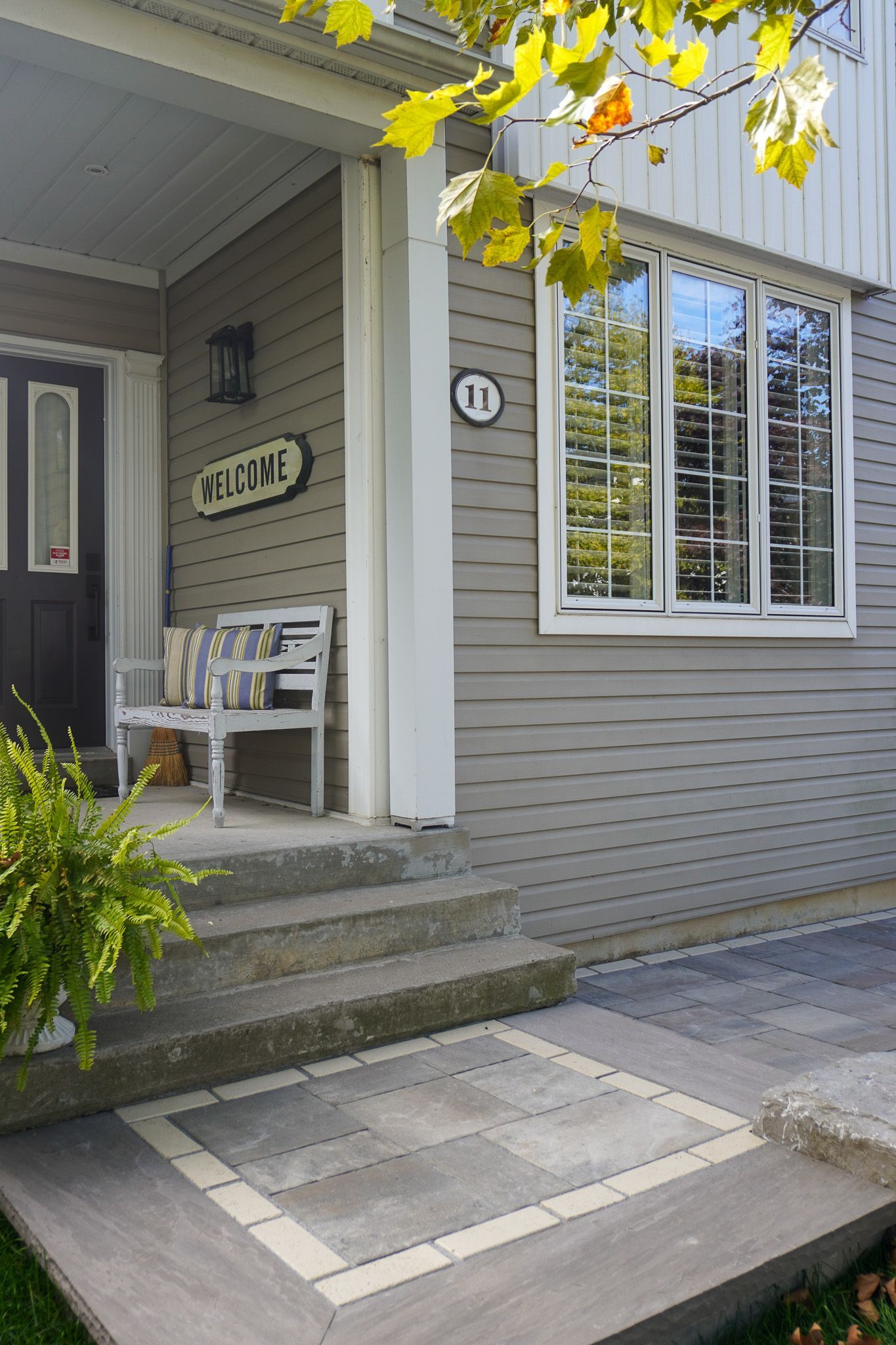 Gray house entrance with steps and a welcome sign. A bench and fern are visible.