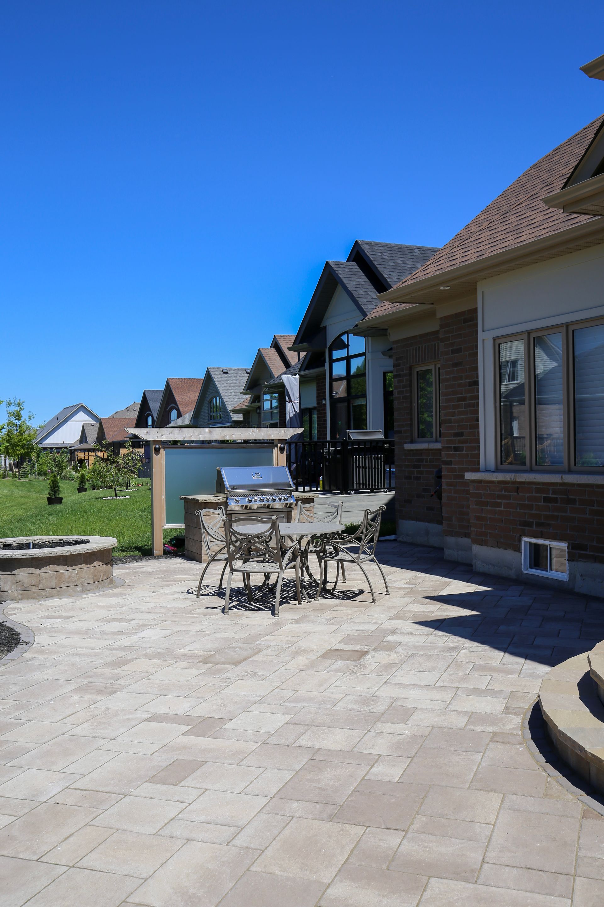 Patio with grill, table, chairs, and surrounding houses under a bright blue sky.