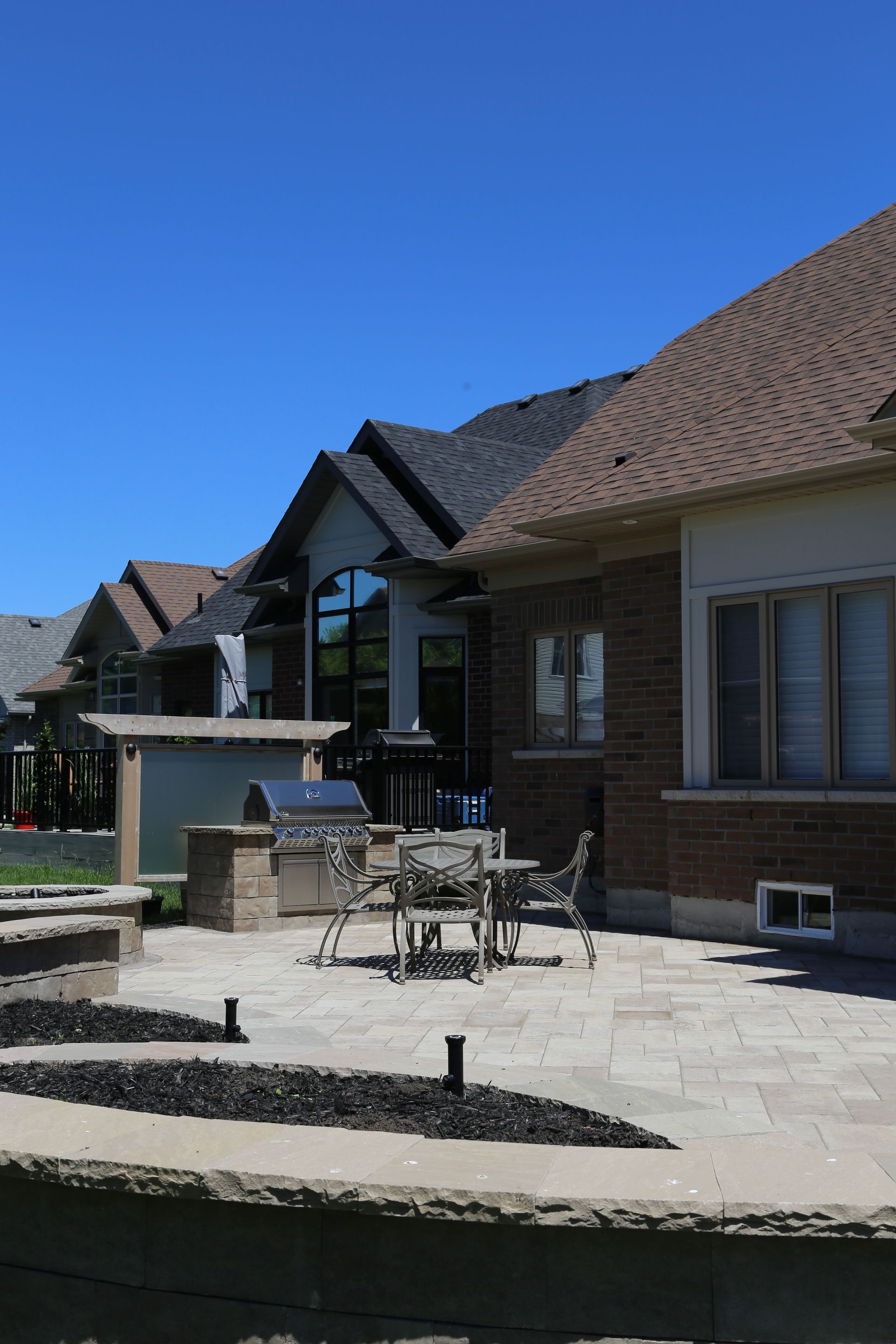 Backyard patio with grill, table, and chairs. Brick houses under a blue sky.