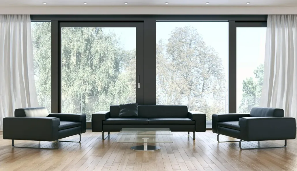 Black leather sofa and armchairs in a modern living room, with large windows overlooking trees and white curtains.