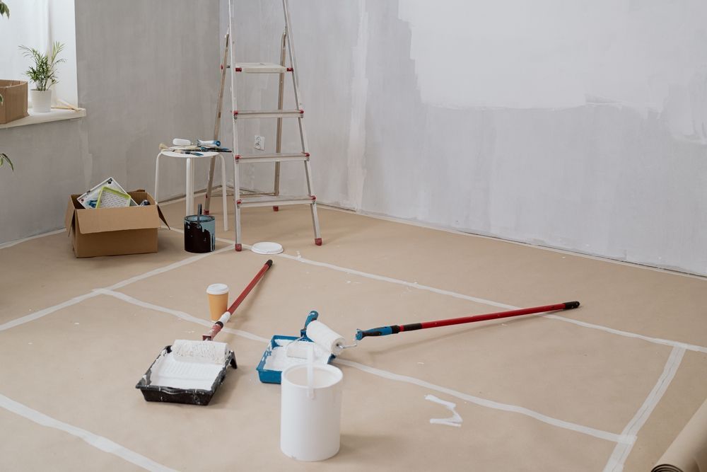 Painting supplies on a paper-covered floor in a room, with a ladder and white-painted wall.