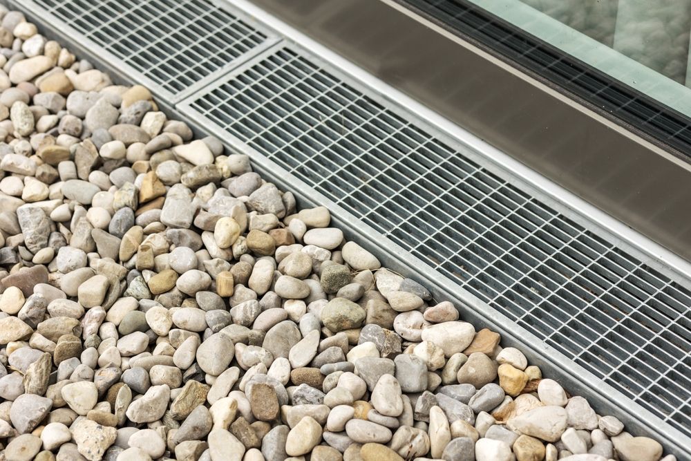 Grey drainage grate beside a bed of smooth white and tan river rocks