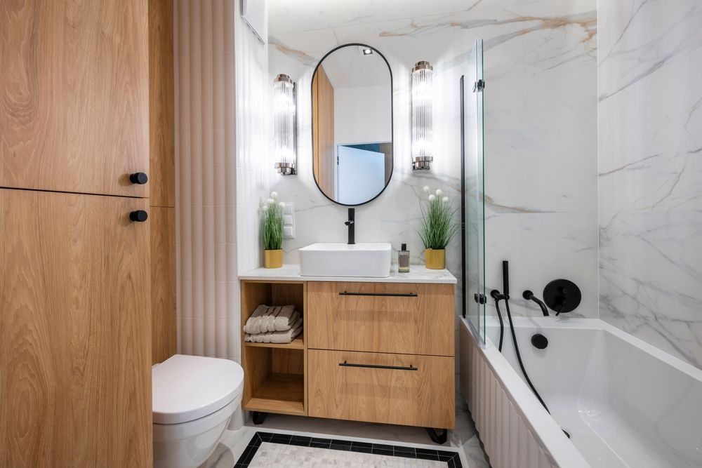 Modern bathroom with wood vanity, oval mirror, white tub, and marble tile walls