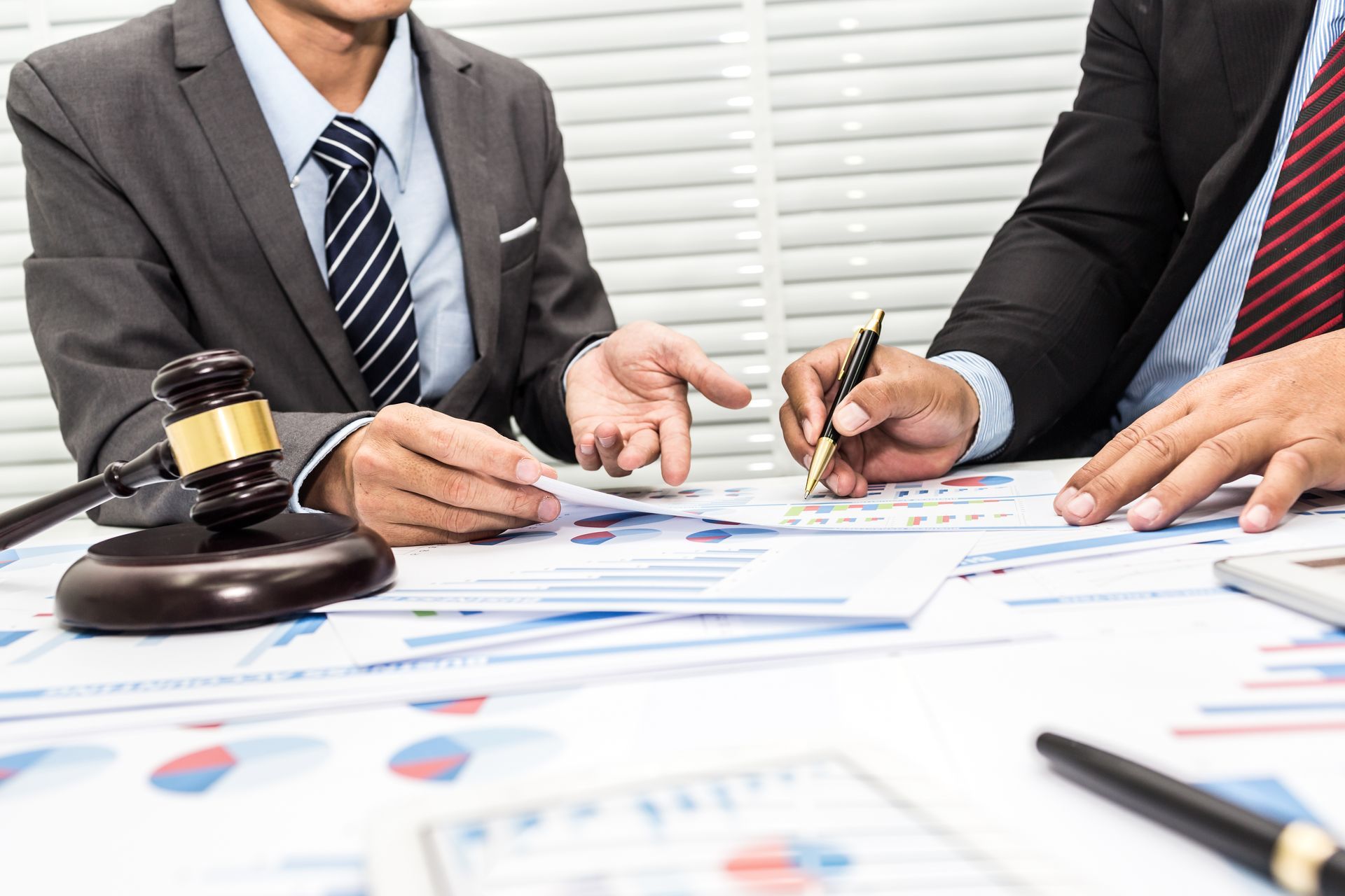 shows two suited professionals reviewing colorful charts and documents beside a wooden judge’s gavel.
