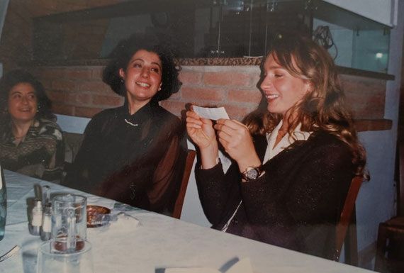 Due donne sedute a un tavolo, sorridenti, si scambiano un piccolo giornale, mentre consumano bevande e piatti in un ristorante scarsamente illuminato.