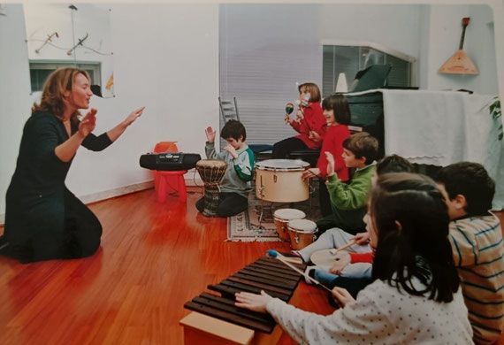 Un'insegnante di musica dirige dei bambini in un'aula con batteria e xilofono su un pavimento di legno.