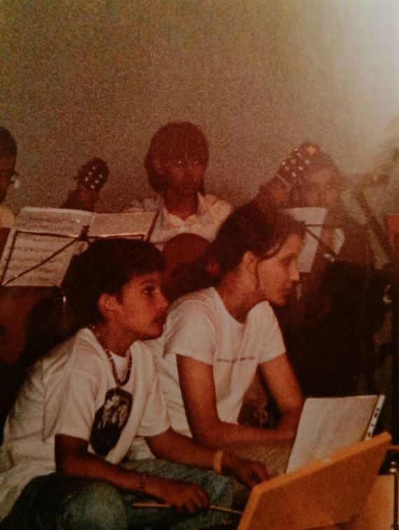Due persone sedute in una sala prove, intente a leggere spartiti musicali, con chitarre e altri musicisti alle loro spalle.