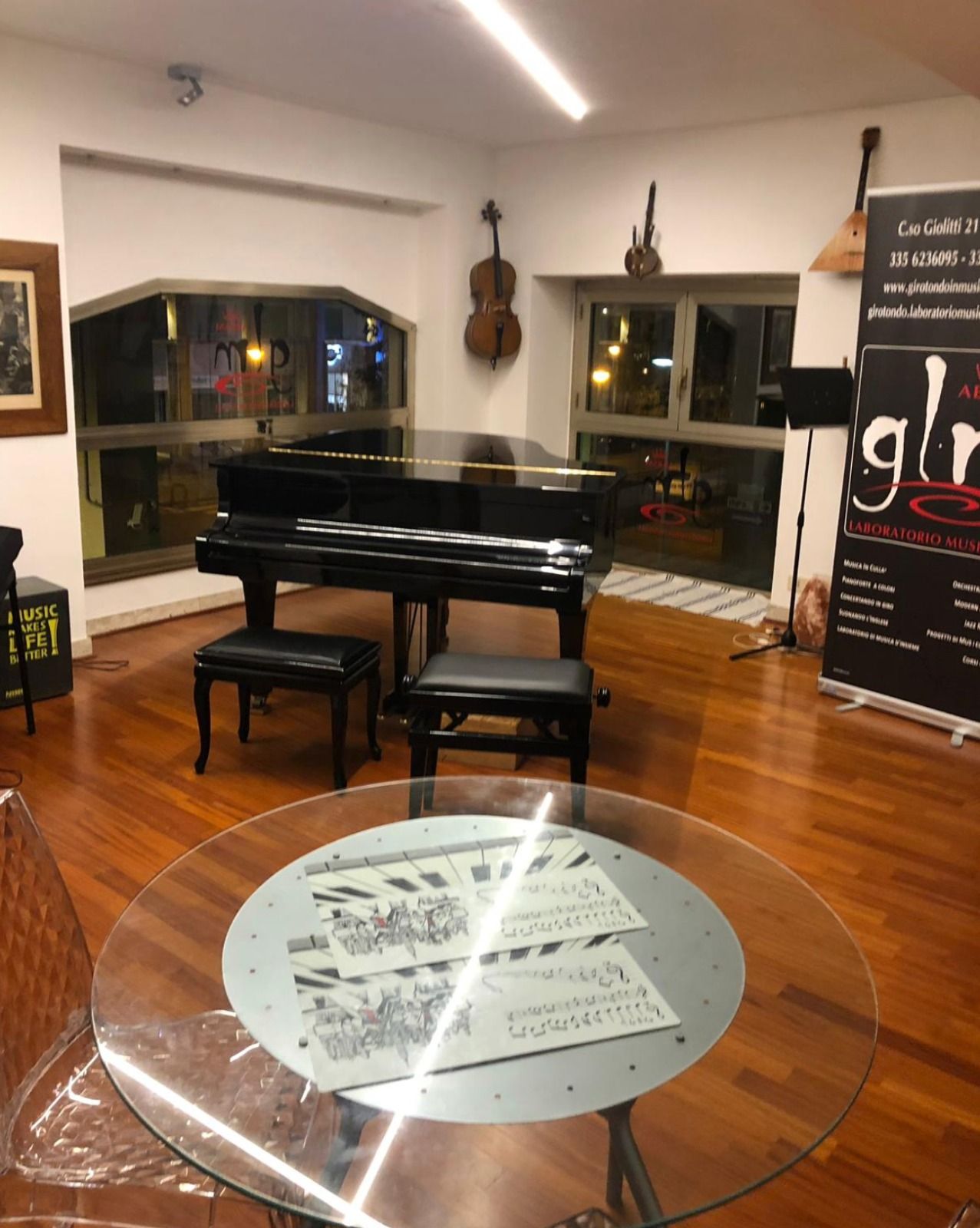 Piccola sala da musica con pianoforte a coda, panca e tavolo rotondo in vetro su pavimento in legno lucido.