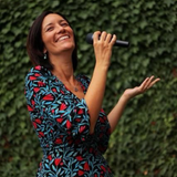 Donna che canta al microfono all’aperto, con un abito a fantasia rossa e blu, sorridente sullo sfondo del verde circostante