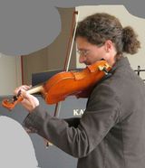 Un musicista suona il violino sul palco, visto di profilo, con un pianoforte sullo sfondo.