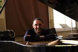 Una persona sorridente appoggiata a un pianoforte a coda in una stanza illuminata dal sole.