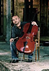 Un uomo seduto con un violoncello in una stanza rustica e poco illuminata, tiene lo strumento in posizione verticale.