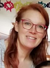 Donna sorridente con capelli rossi e occhiali rosa, seduta in un ambiente interno contro una parete decorata.