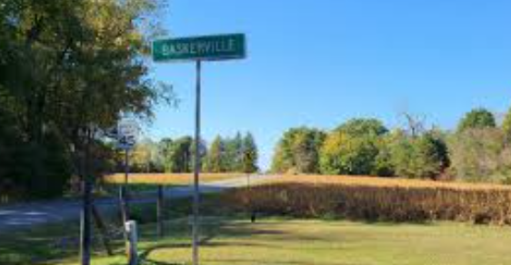 A green street sign labeled BASKERVILLE stands on a metal pole in a rural, sunny setting with trees and a field. A green street sign labeled BASKERVILLE stands on a metal pole in a rural, sunny setting with trees and a field.