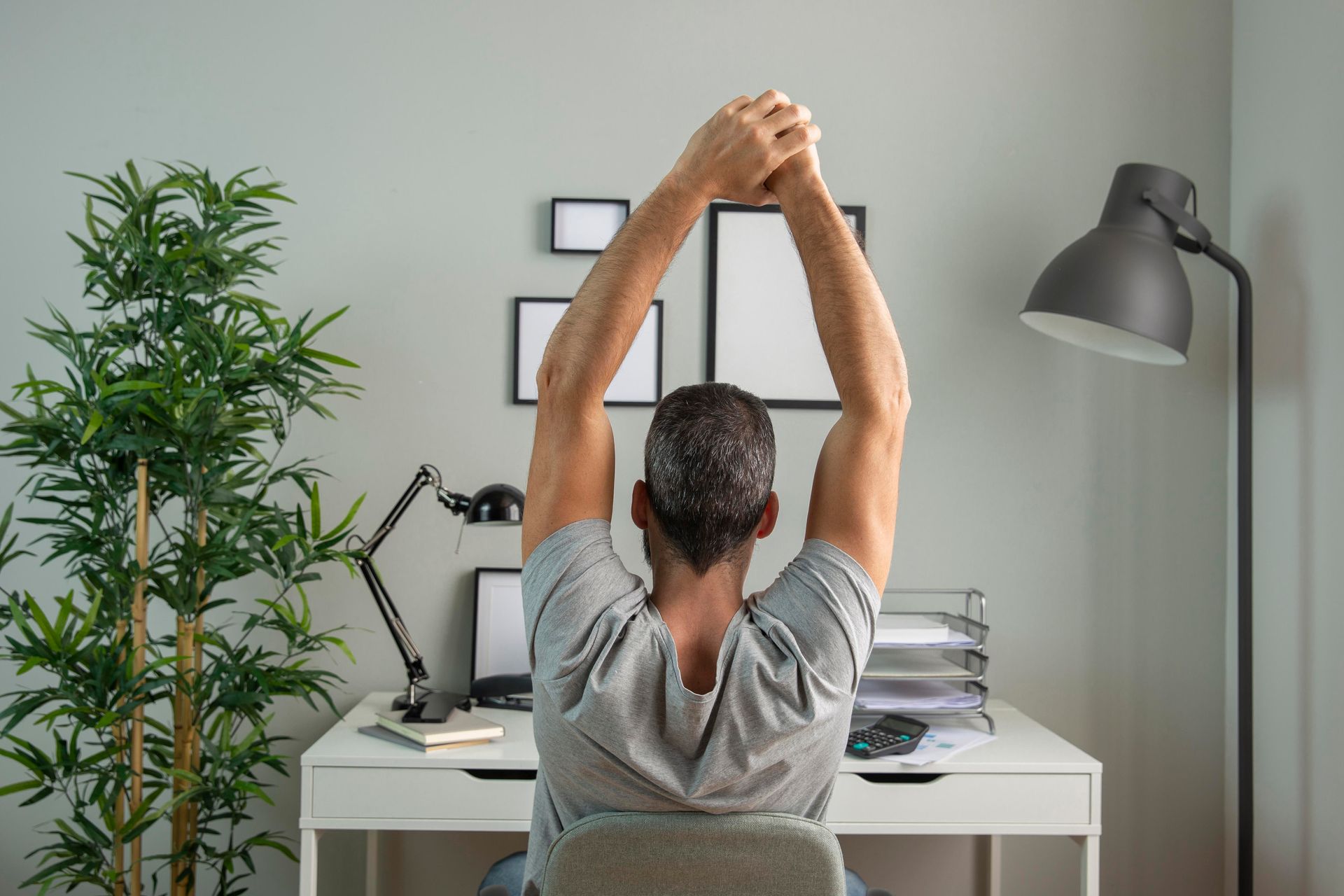 Person stretching with arms raised at a white desk in a home office, seen from behind.