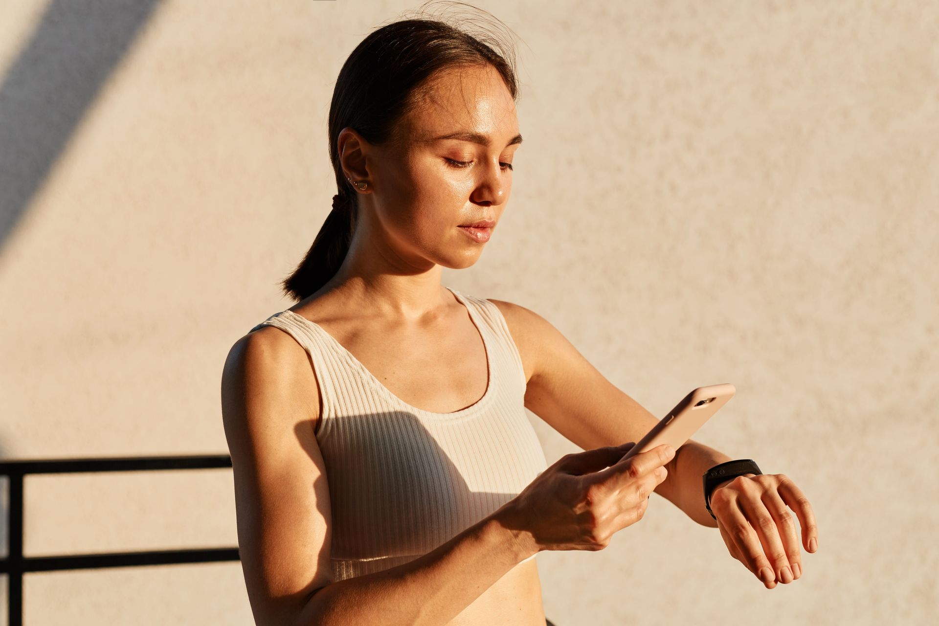 Woman in a tank top checking her smartwatch while holding a smartphone on a sunlit balcony