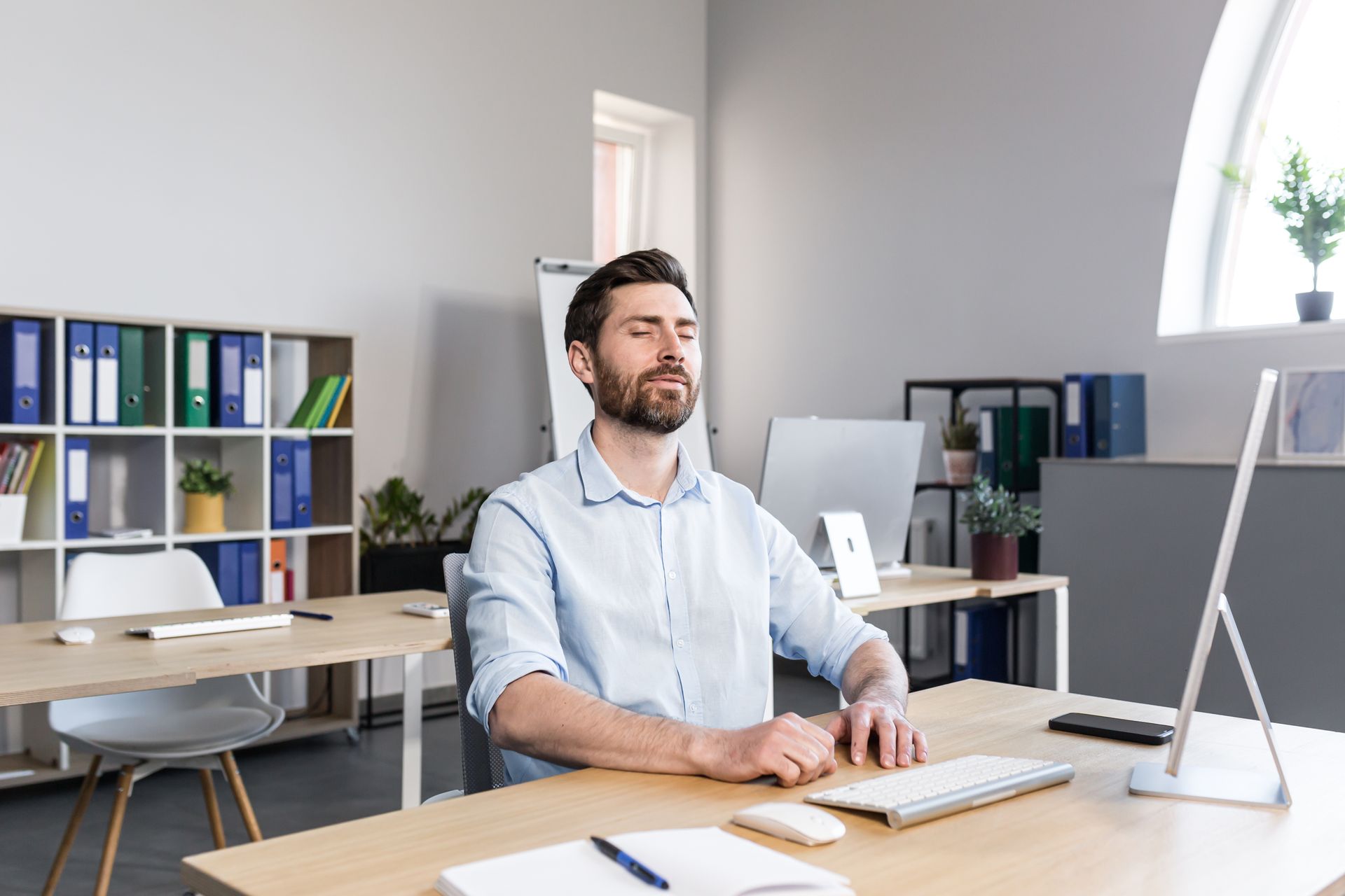 Man working at a desk in a bright office, typing on a laptop with shelves and files behind him