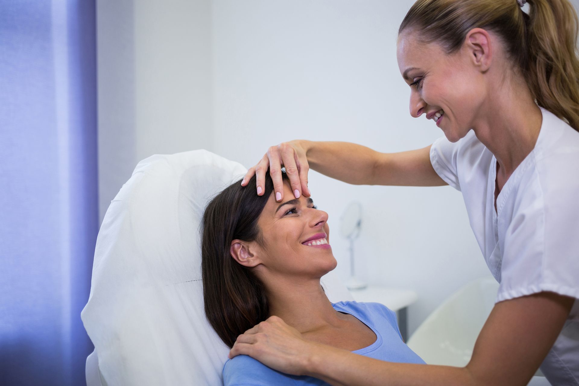 Nurse smiling and checking a relaxed patient’s forehead in a bright clinic room