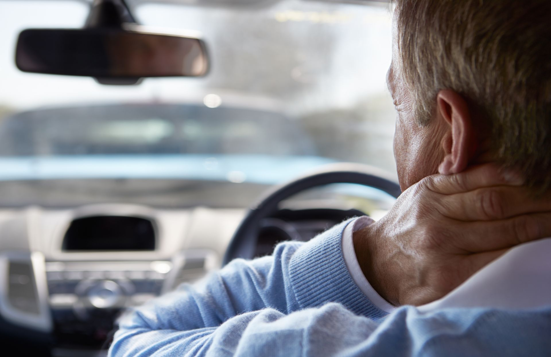 Driver rubbing neck while sitting in a car, seen from behind with dashboard and steering wheel visible