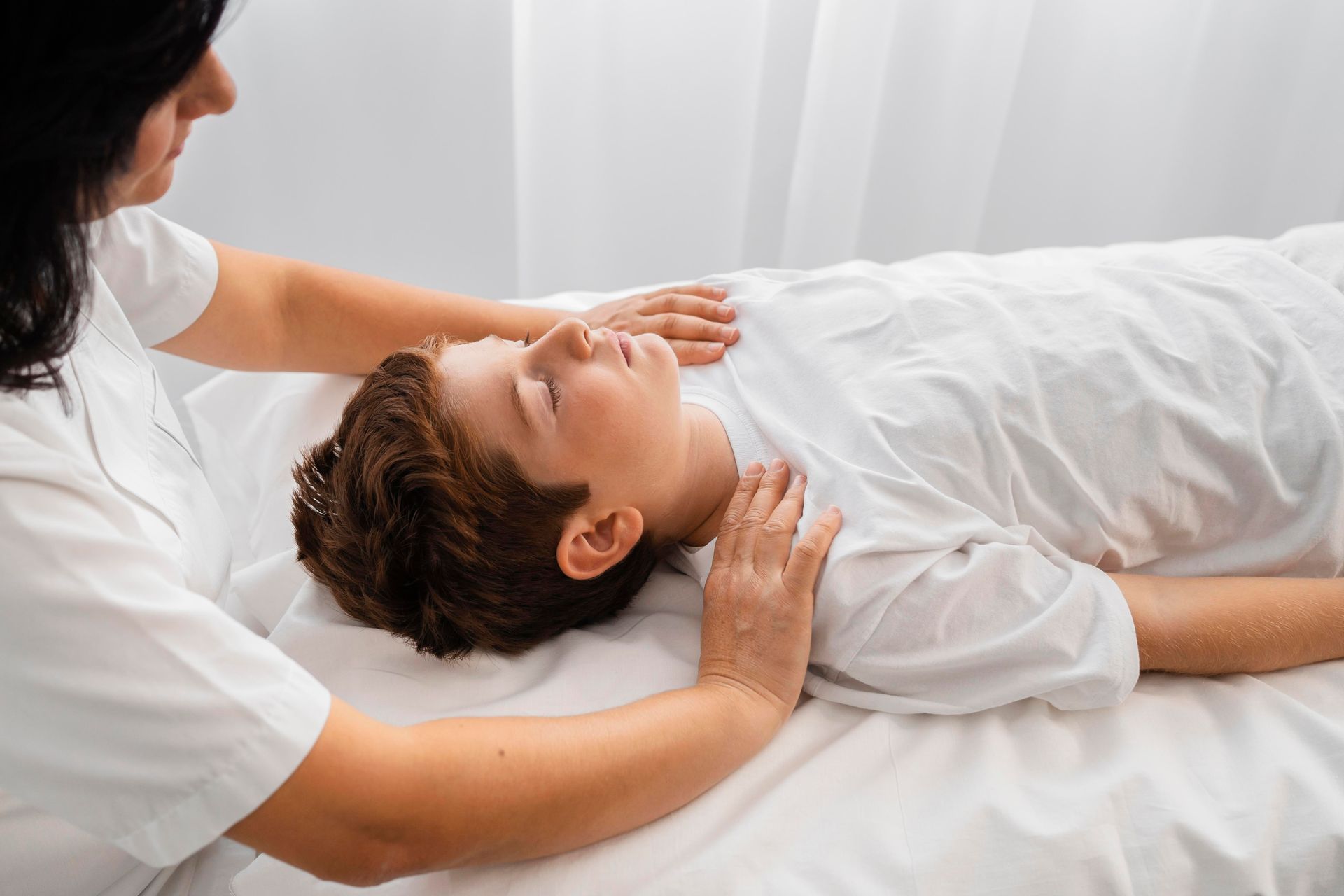 Therapist gently performs a head and shoulder massage on a reclining client in a white treatment room