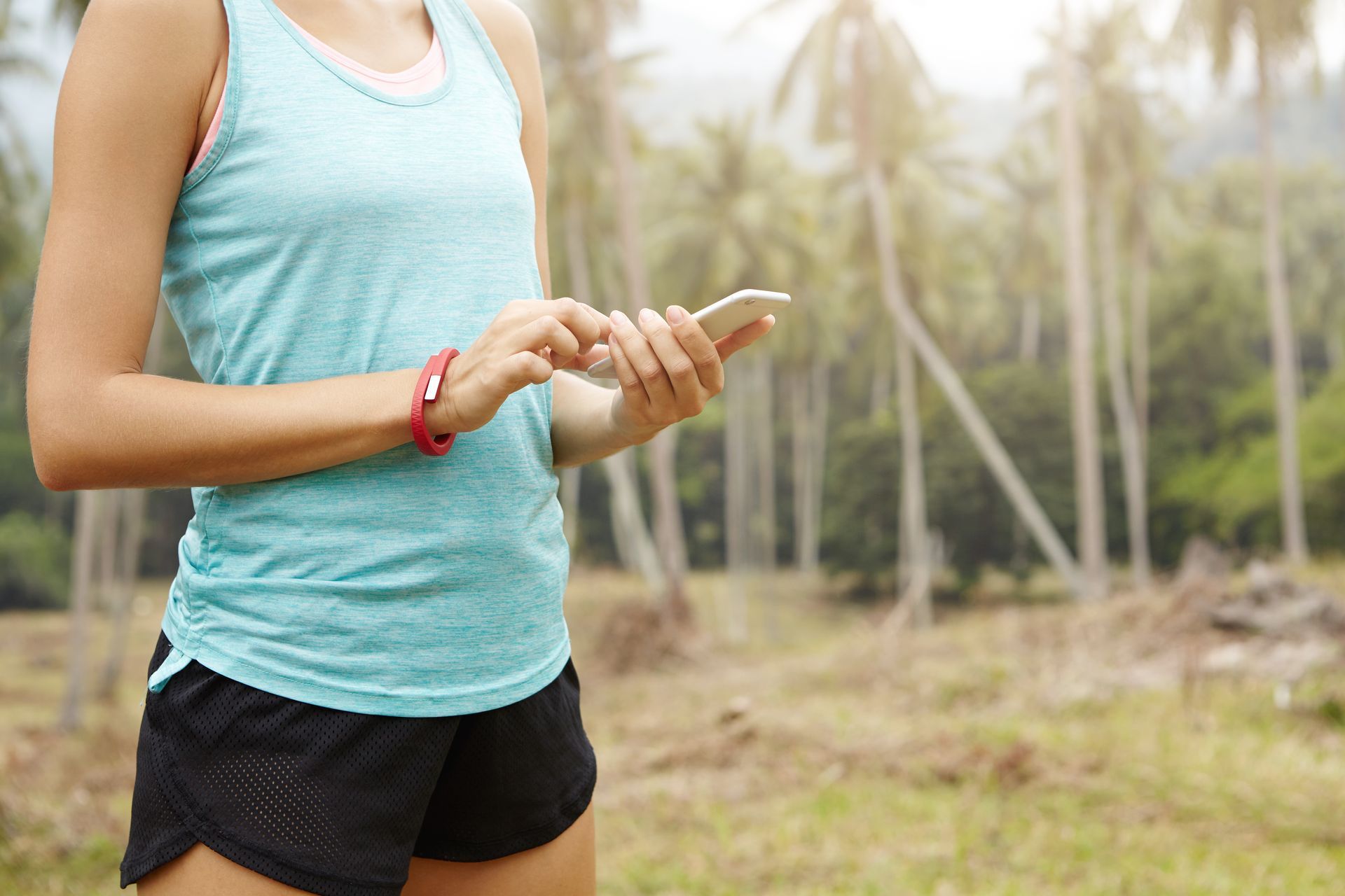 Person in teal tank top and black shorts holding a phone outdoors near palm trees.