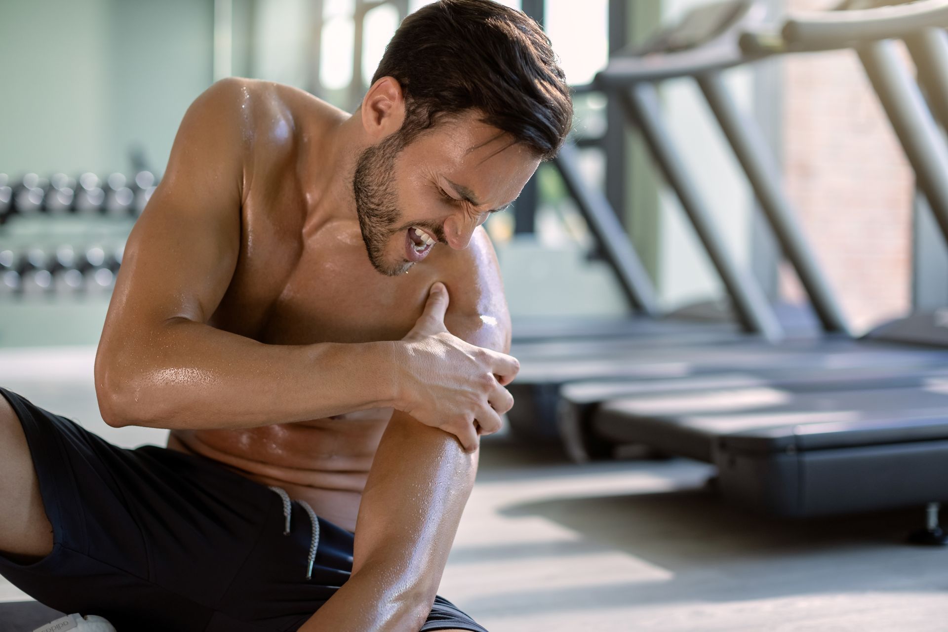 Man in a gym, leaning forward and holding his shoulder, appearing to feel pain or strain.