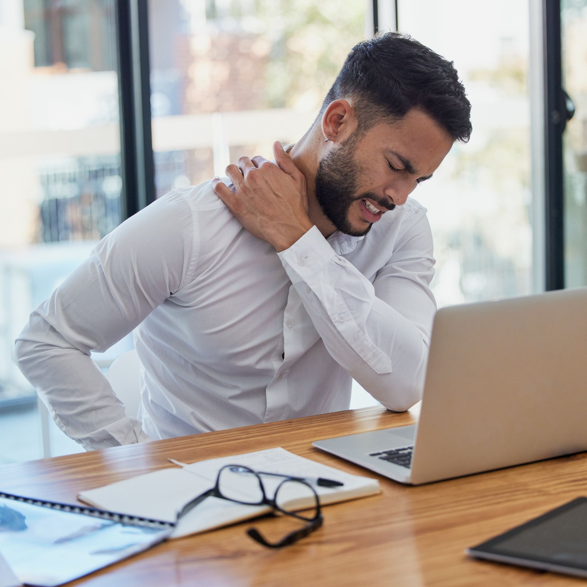 Man holding his neck at a desk beside a laptop, looking pained or tense