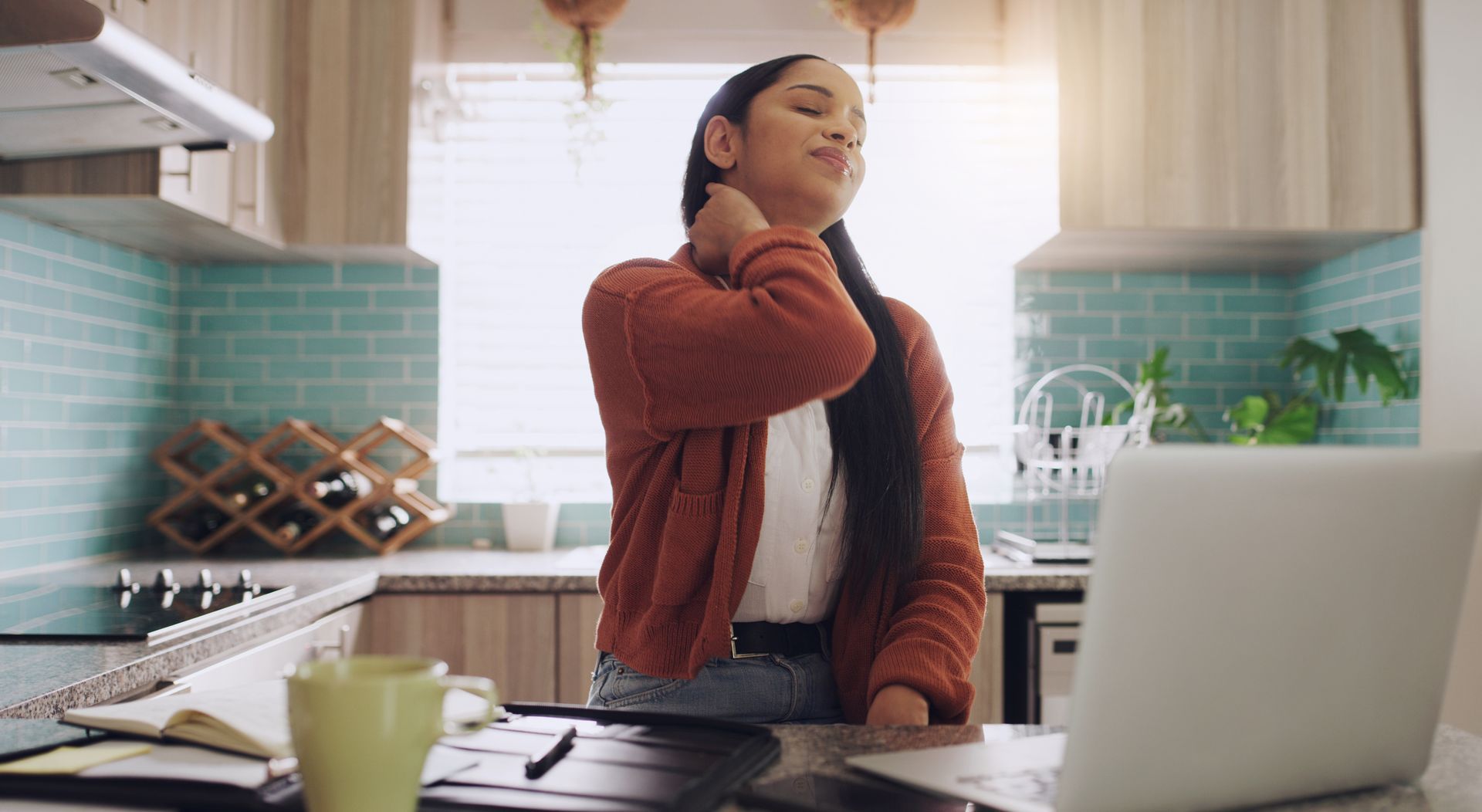 Woman stretching at a kitchen table with a laptop, wearing a red sweater in a bright home kitchen.