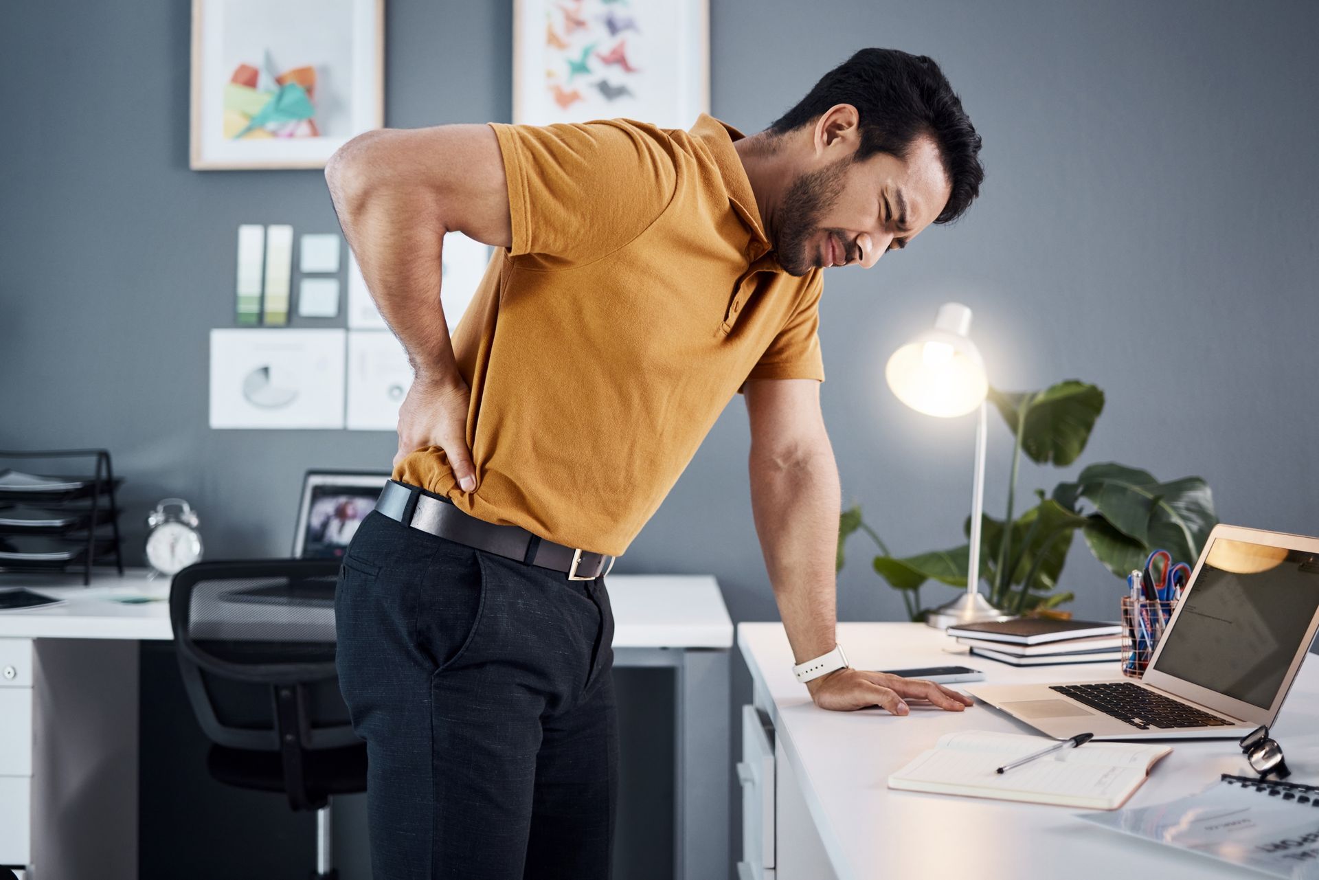 Man leaning over a desk and holding his lower back in an office, with a laptop and desk lamp nearby