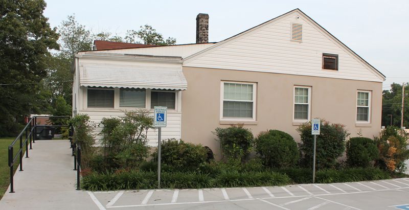 Small beige building with a ramp, shrubs, and accessible parking signs in front