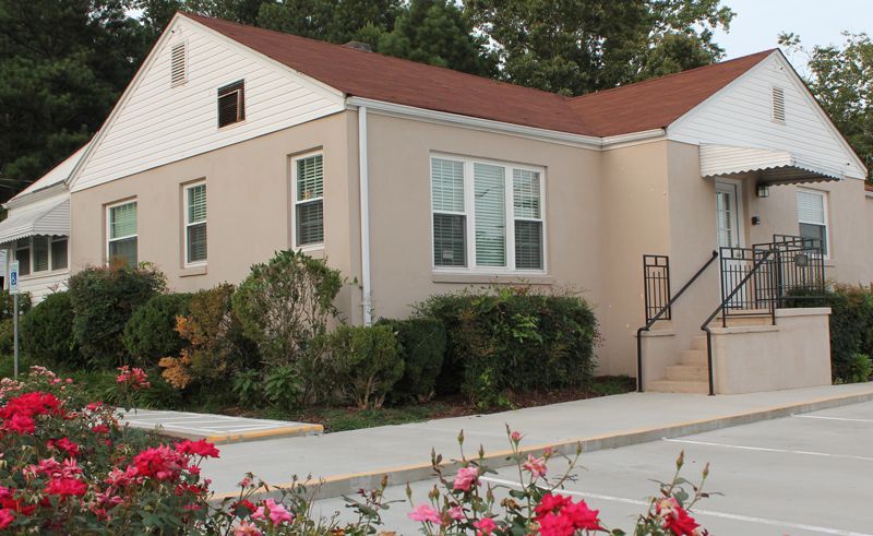 Small beige building with red roof, white shutters, and steps at the entrance, beside a parking lot and flowers