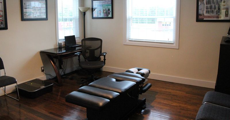 Small massage room with a black table, desk, chair, and wall art by two windows.