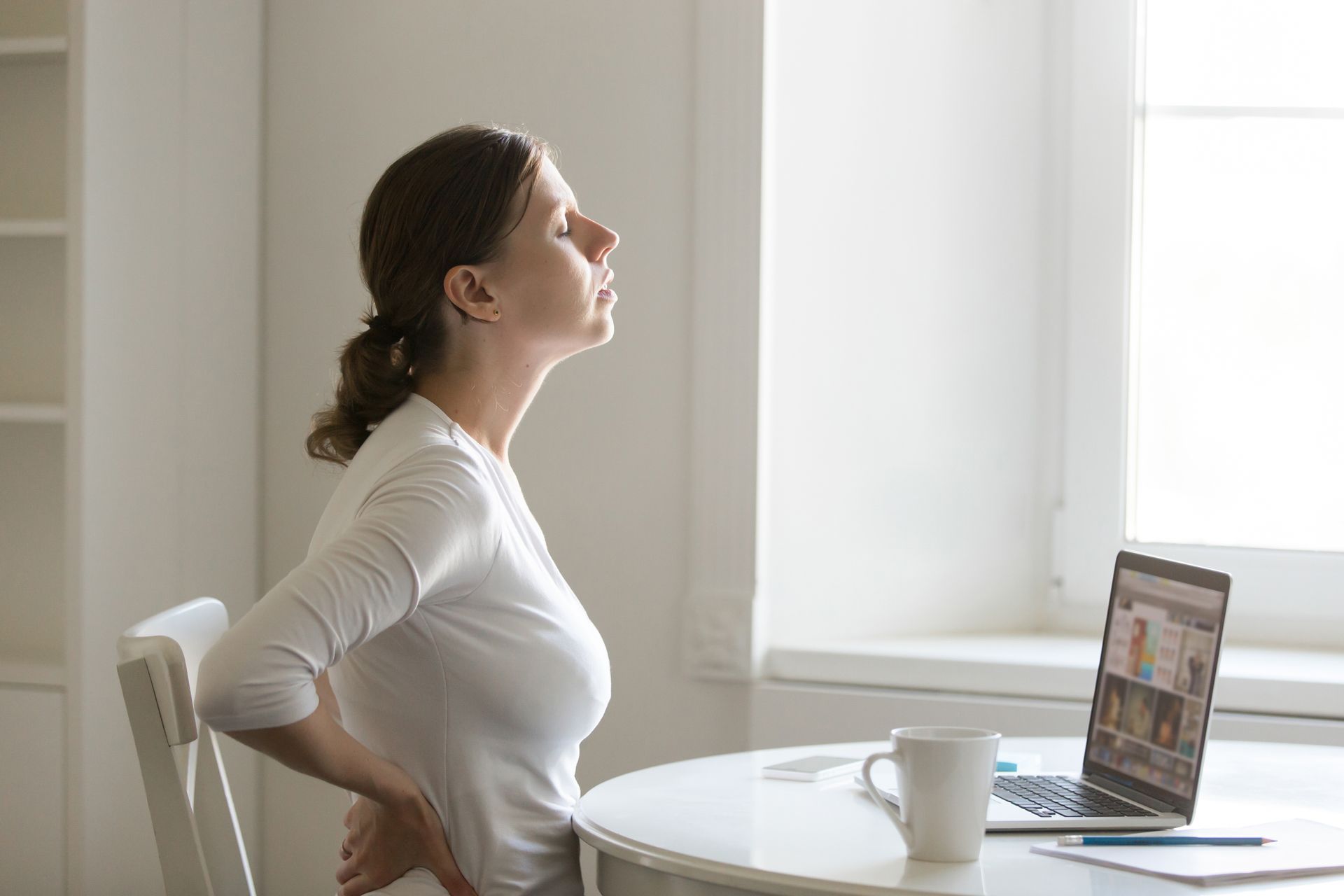 Woman sitting at a desk, stretching her back while looking toward a laptop near a bright window.