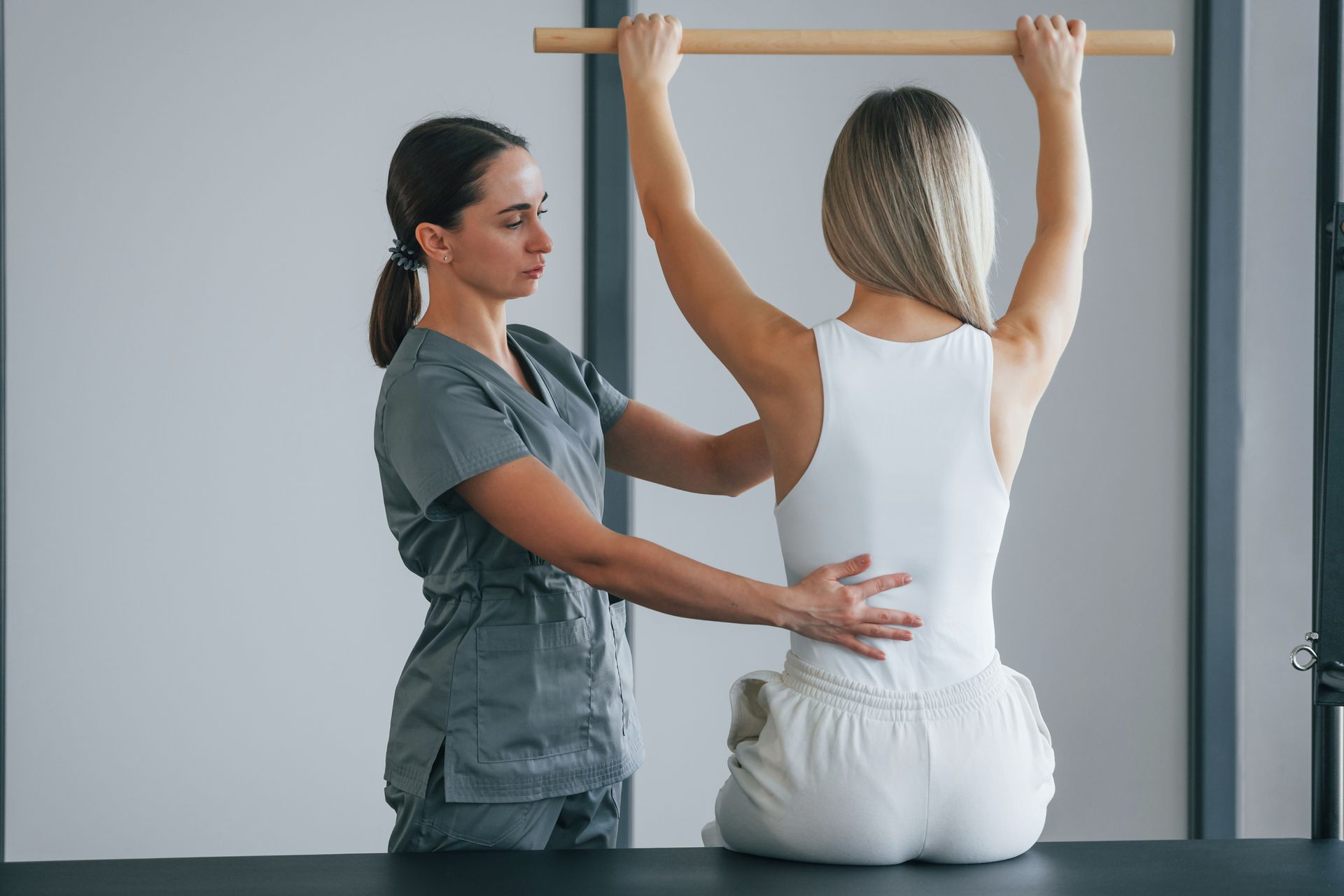 Physical therapist assisting a seated patient with an arm stretch on a wall-mounted bar in a clinic room