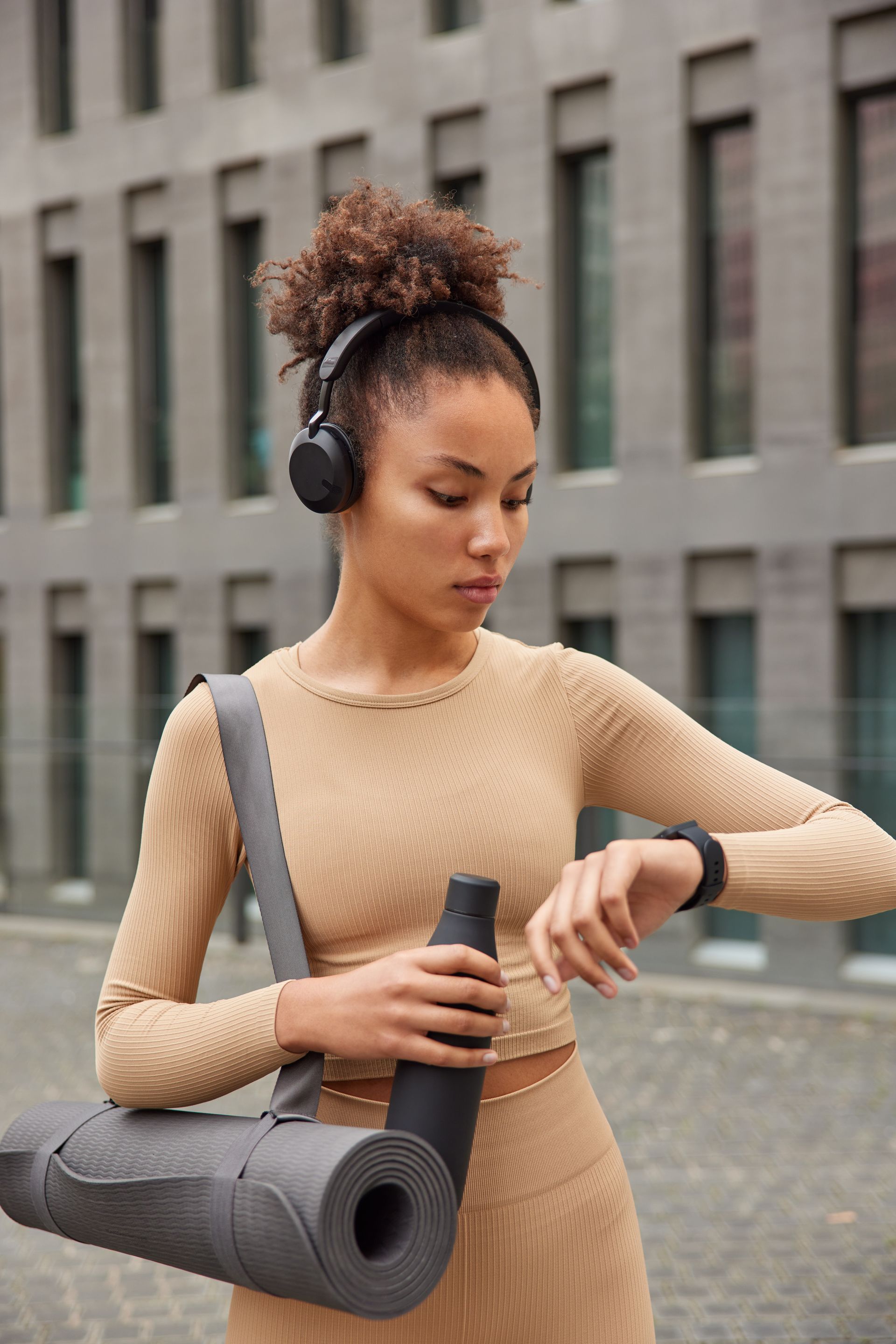 Woman in beige athletic wear holding a yoga mat and water bottle outdoors, wearing headphones.
