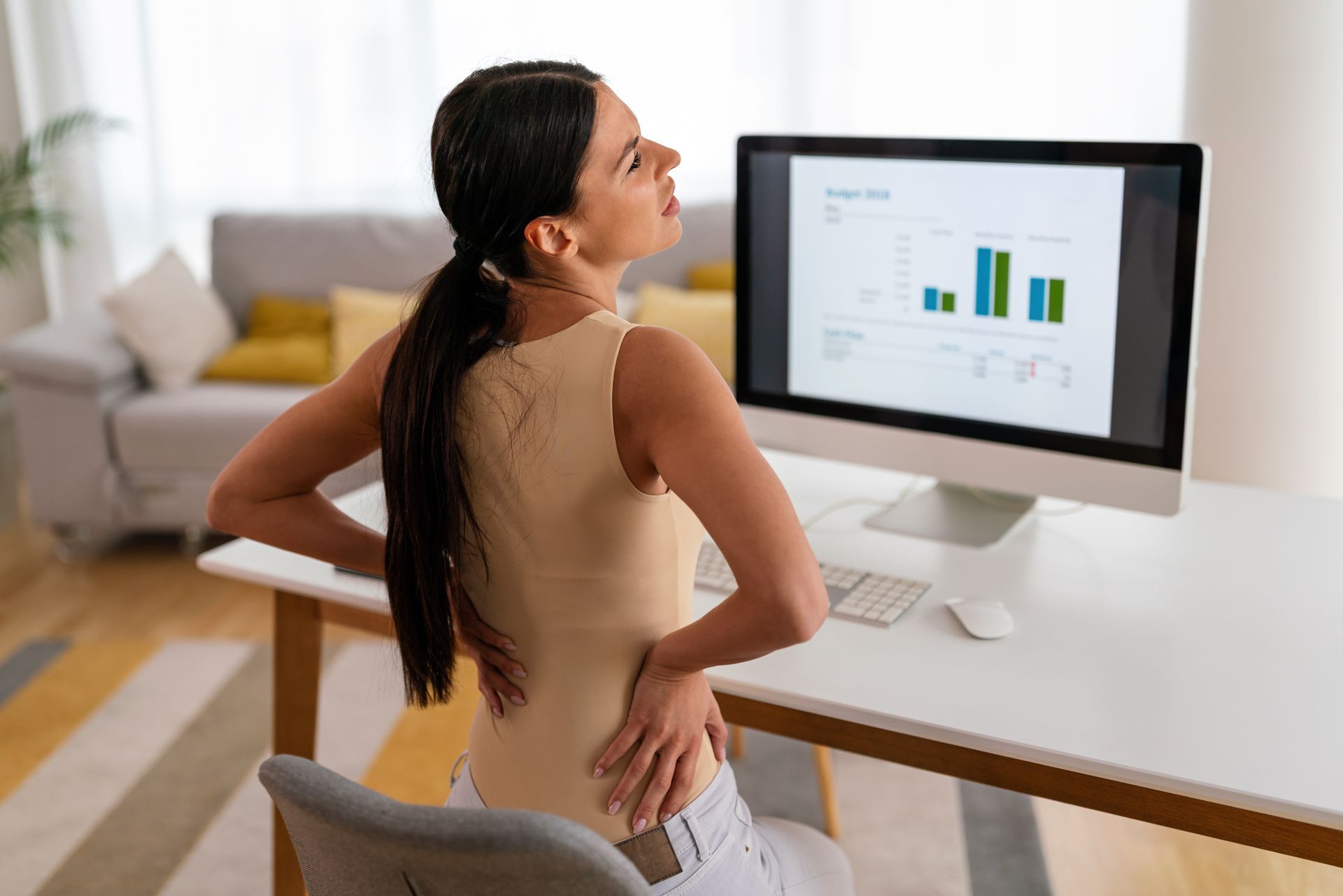 Woman holding her lower back while viewing charts on a desktop computer at a desk