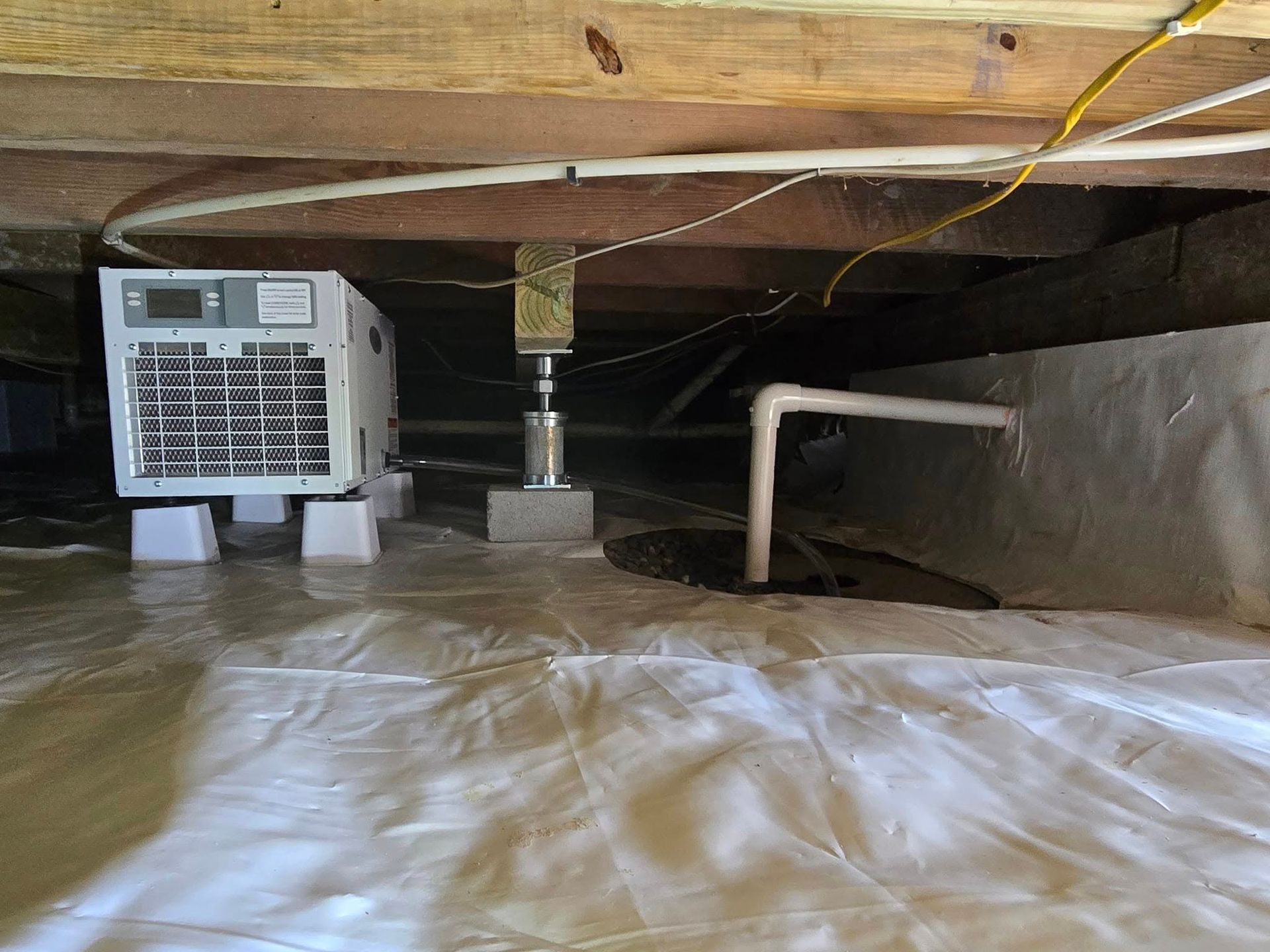 Crawl space with a dehumidifier, plastic floor barrier, exposed beams, and pipes.
